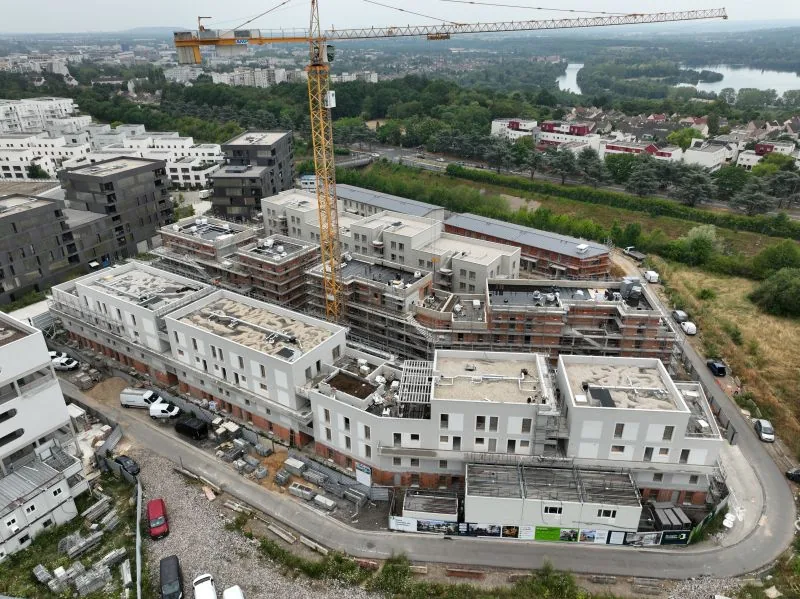 Drone construction site monitoring — aerial view of residential buildings under construction around a tower crane, with the river in the background, Dronexperts